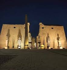 Vista noturna do Templo de Luxor iluminado, com colunas monumentais e obelisco na entrada
